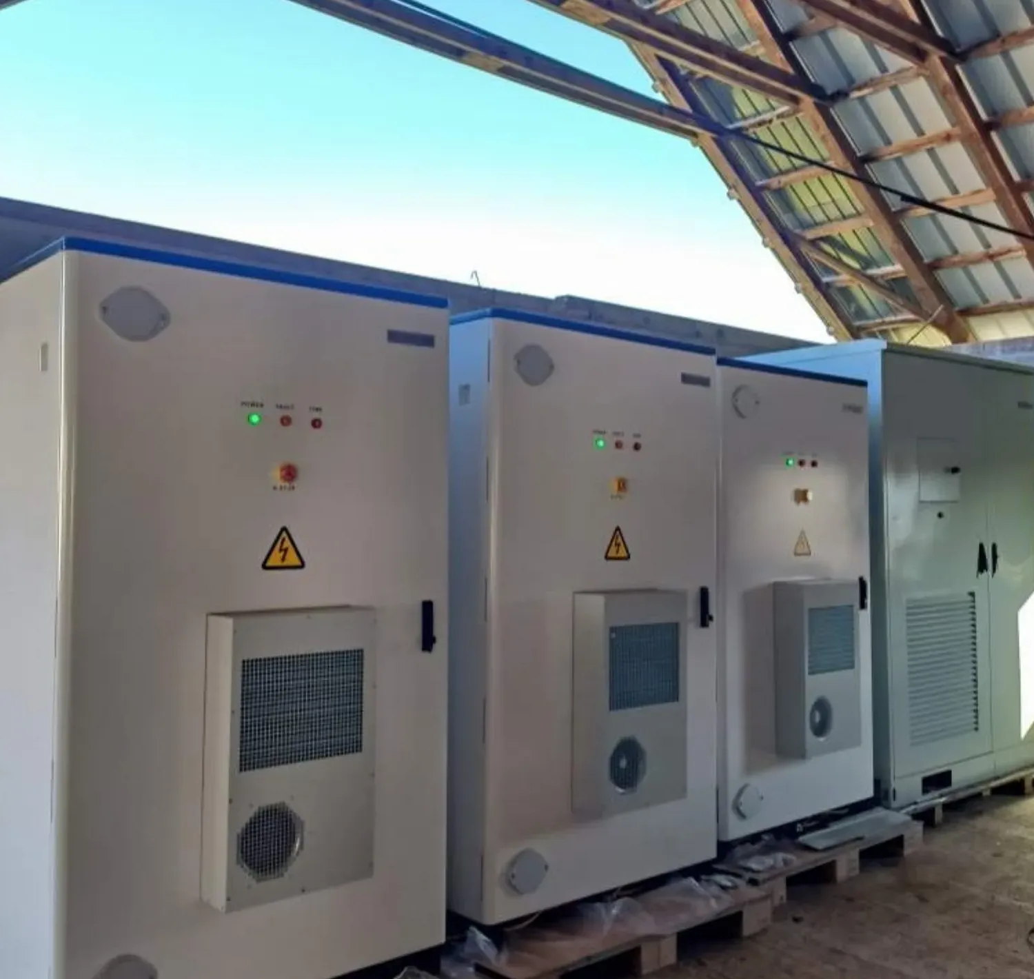 Three air-cooled battery storage cabinets lined up under a roof before installation, cooling units and status indicators on their front doors
