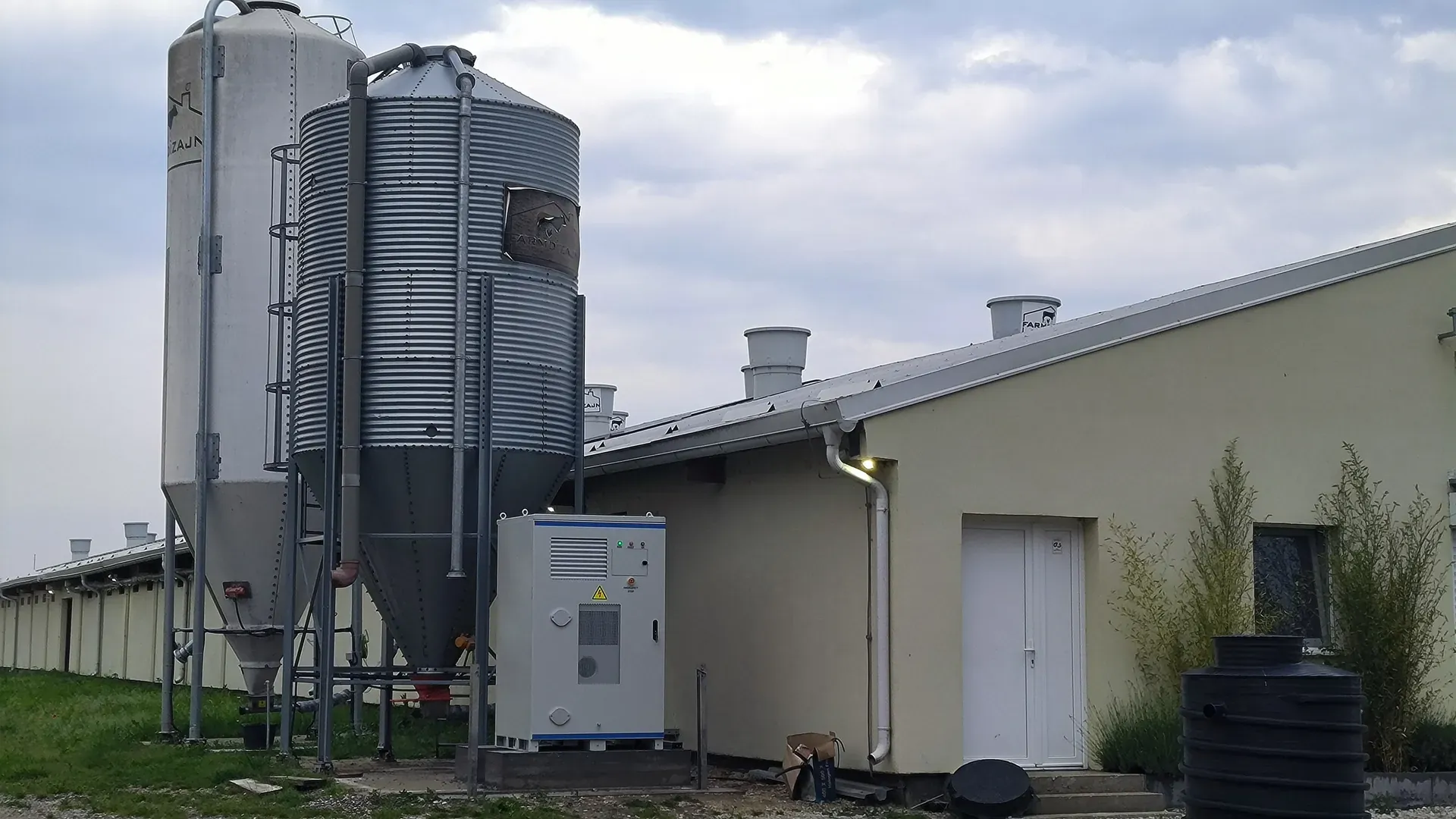 Outdoor battery storage cabinet on a concrete pad at a farm site, with grain silos and a farm building behind it
