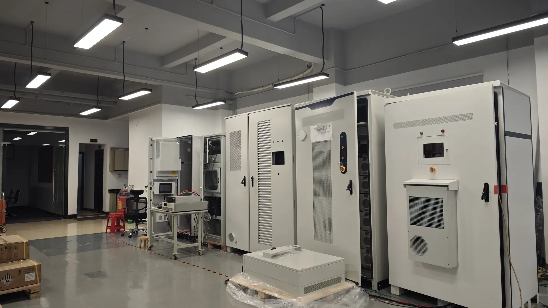 White battery storage cabinets on the floor of a testing hall with a wheeled instrument bench alongside, ahead of factory acceptance testing