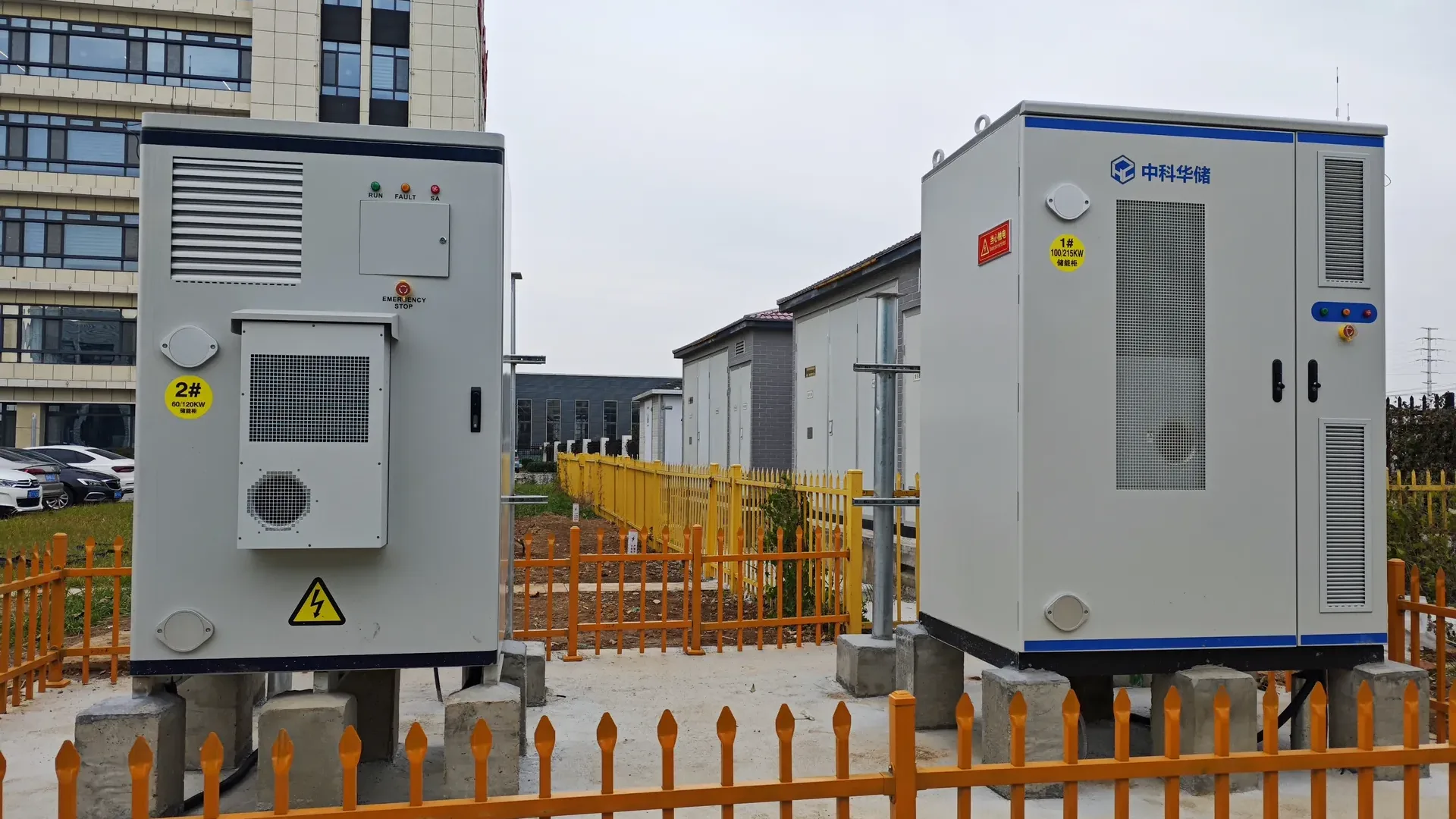 Two outdoor battery storage cabinets in a fenced compound on an industrial project site, seen during commissioning