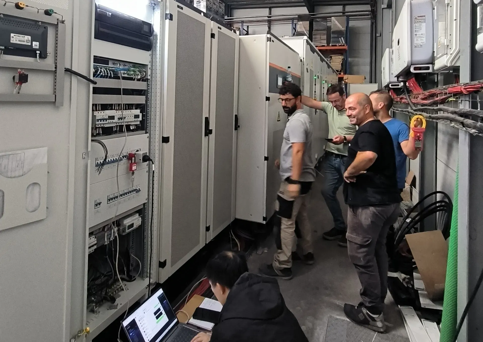 Engineers at a row of storage and control cabinets in a workshop, with monitoring software open on a laptop in the foreground